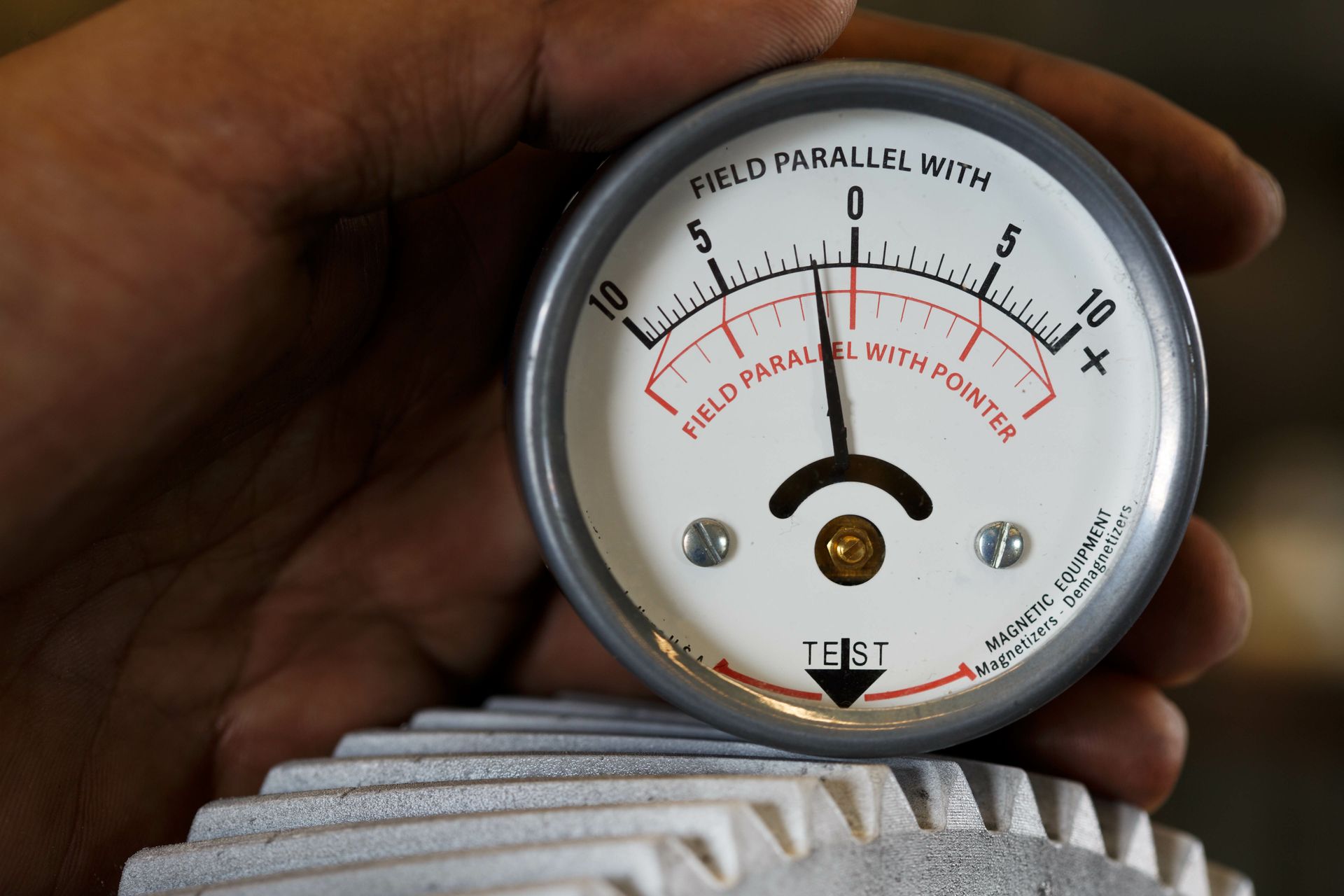 A hand holding a magnetic field meter, reading about 3. The meter sits on a ridged metal surface.