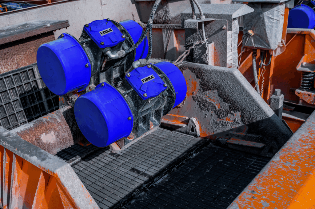Two blue industrial vibratory motors on a metal machine sorting material, inside a colorful orange hopper.